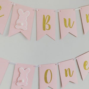 Op de afbeelding: Roze en gouden banner met de woorden "Bunny One" in gouden glitterletters. De banner heeft roze konijnenuitsnijdingen met witte staarten.