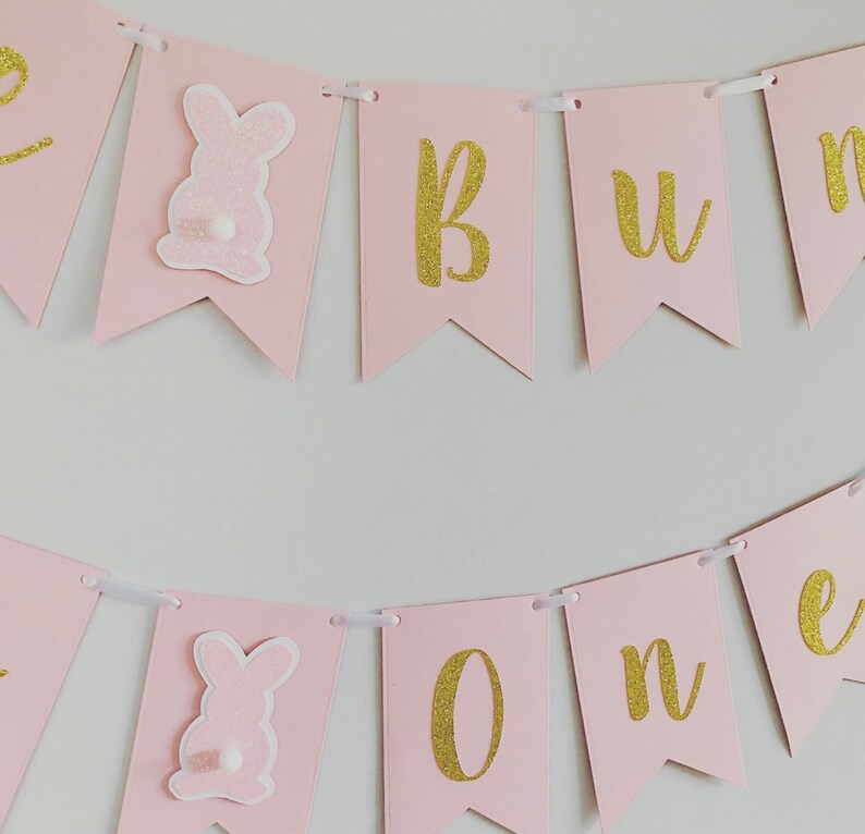 Op de afbeelding: Roze en gouden banner met de woorden "Bunny One" in gouden glitterletters. De banner heeft roze konijnenuitsnijdingen met witte staarten.