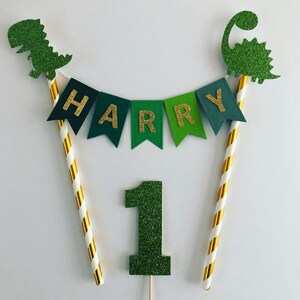 May include: Green and gold glitter cake topper with the name "HARRY" in gold letters on a banner, the number "1" and two dinosaur figures.