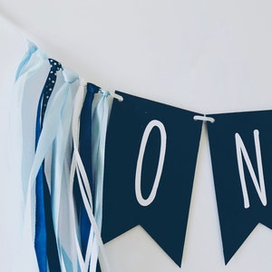 Puede incluir: Una pancarta azul y blanca con la palabra "ONE" en letras blancas sobre banderines negros. La pancarta tiene cintas azules y blancas.