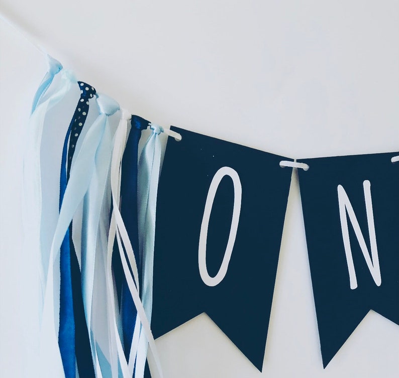 Puede incluir: Una pancarta azul y blanca con la palabra "ONE" en letras blancas sobre banderines negros. La pancarta tiene cintas azules y blancas.
