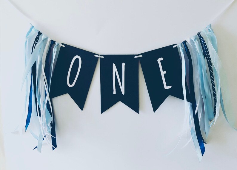 Puede incluir: Una pancarta azul marino con letras blancas que deletrean "ONE" con cintas azules y blancas colgando de cada extremo.