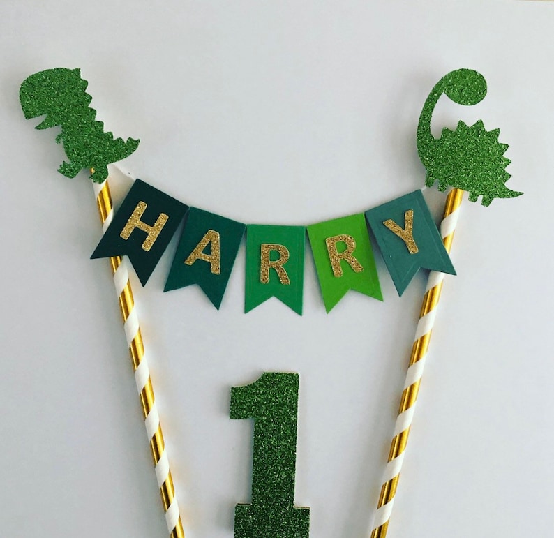 May include: Green and gold glitter dinosaur cake topper with a banner that says "HARRY" and a green glitter number one.