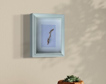 May include: A light blue picture frame with a white mat and a dried twig inside. The frame is hanging on a light beige wall. A geometric planter with a succulent plant is on a wooden table.