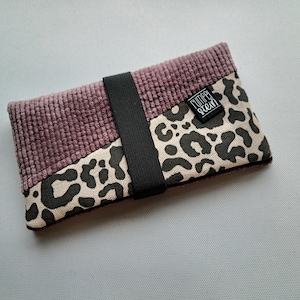 Könnte beinhalten: Ein kleines, rechteckiges Portemonnaie mit einer Kordstruktur in einem gedämpften Lila. Das Portemonnaie hat einen Abschnitt mit einem schwarzen und beigefarbenen Leopardenmuster und ein schwarzes Gummiband. Das Etikett lautet "CUMPER STEIN".
