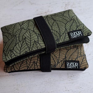 Könnte beinhalten: Zwei grüne und braune Stoffbeutel mit schwarzen Riemen und einem Blatt-Muster. Die Beutel haben ein schwarzes Etikett mit dem Text "Rumpel Stiem" darauf.