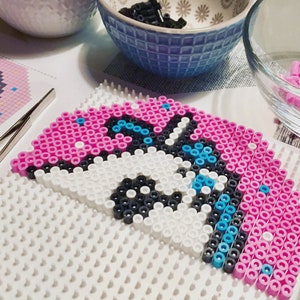 Puede incluir: Primer plano de un diseño de arte de píxeles de un unicornio parcialmente completado en una placa de clavijas blanca. El unicornio está hecho de cuentas de plástico rosa, blanco, negro y azul.