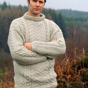 May include: A man wearing a light grey cable knit jumper with a chevron pattern. He is standing in front of a blurred background of trees and undergrowth.