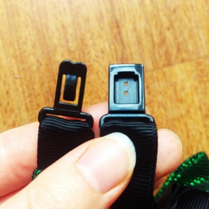 May include: Close-up of two black adjustable straps with plastic buckles. The buckles have two small holes. The straps are attached to a green and black ribbon.