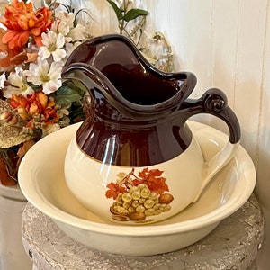 May include: A brown and white ceramic pitcher with a grape and leaf design sits inside a white ceramic bowl. The pitcher and bowl are on a weathered, gray wooden stool.