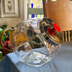 May include: A clear glass jar with a silver lid and a red knob. The jar has a unique, rounded shape with a wide opening and a ribbed design. The jar is sitting on a blue surface.