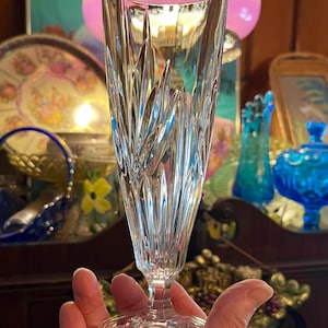 Peut inclure: Un fl&ucirc;te &agrave; champagne en cristal transparent avec un motif vertical textur&eacute;. Le verre a une longue tige et un dessus &eacute;vas&eacute;. La base du verre a un autocollant dor&eacute;. L'arri&egrave;re-plan comprend divers objets d&eacute;coratifs.
