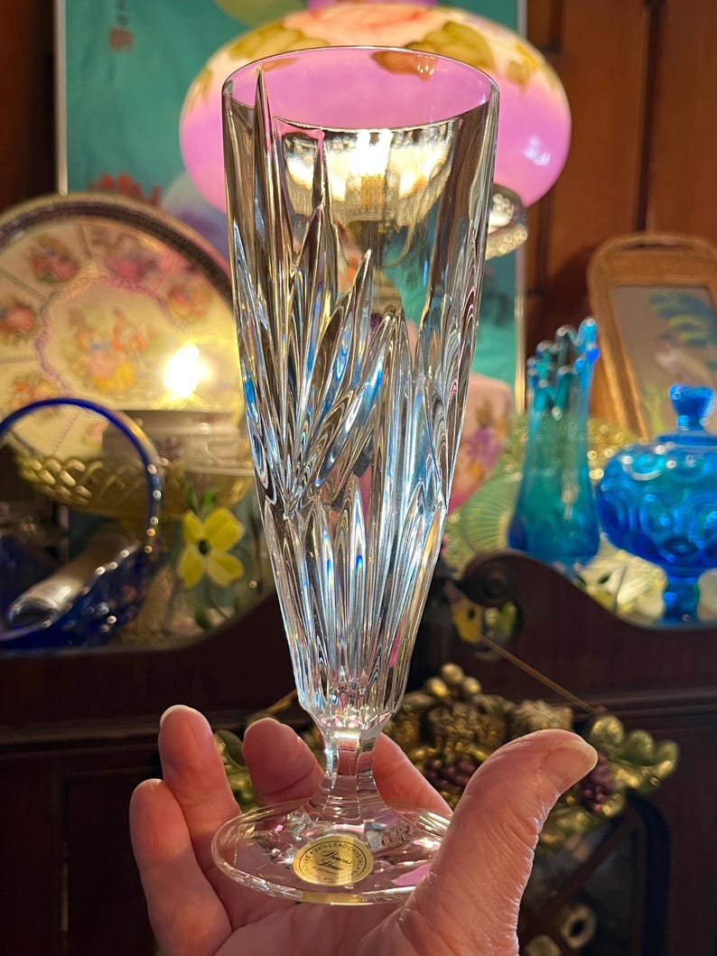 Peut inclure: Un fl&ucirc;te &agrave; champagne en cristal transparent avec un motif vertical textur&eacute;. Le verre a une longue tige et un dessus &eacute;vas&eacute;. La base du verre a un autocollant dor&eacute;. L'arri&egrave;re-plan comprend divers objets d&eacute;coratifs.