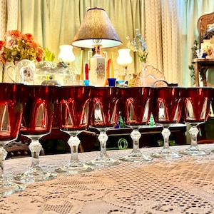 May include: A collection of eight ruby red wine glasses with a cut glass design. The glasses are arranged on a white lace tablecloth, with a lamp and antique furniture in the background.