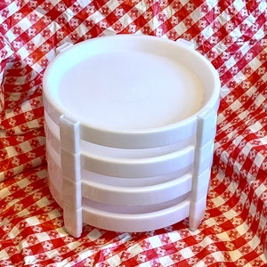 May include: A stack of four white plastic serving trays with a rounded top and a series of horizontal ridges. The trays are supported by vertical white supports. The background is a red and white checkered pattern.