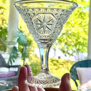 May include: A clear, vintage-style glass with a detailed, textured pattern. The bowl has a starburst design at the center, surrounded by a geometric pattern. The stem is slender, and the base is also patterned. The glass is held up against a blurred green background.