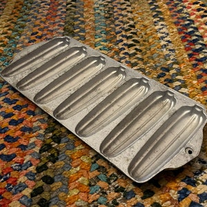 May include: A vintage aluminum baking pan with eight long, narrow slots for making cornbread or other baked goods. The pan has a handle on one side.