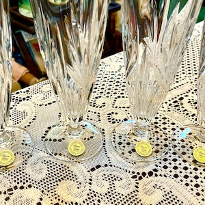 Peut inclure: Quatre fl&ucirc;tes &agrave; champagne en cristal transparent avec des coupes verticales et diagonales complexes. Chaque fl&ucirc;te a une base ronde et un autocollant dor&eacute; avec du texte. Sur une nappe en dentelle blanche.