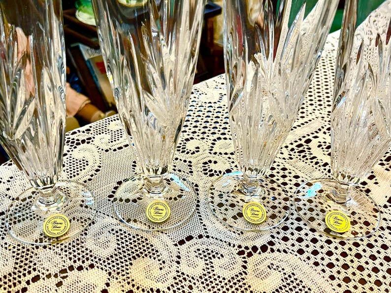 Peut inclure: Quatre fl&ucirc;tes &agrave; champagne en cristal transparent avec des coupes verticales et diagonales complexes. Chaque fl&ucirc;te a une base ronde et un autocollant dor&eacute; avec du texte. Sur une nappe en dentelle blanche.