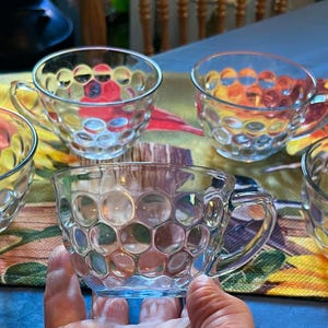 May include: A set of five clear glass teacups featuring a raised honeycomb or bubble pattern on the exterior surface. Each cup includes a small handle on the side and a wide, circular opening, showcasing a transparent design with a textured finish.