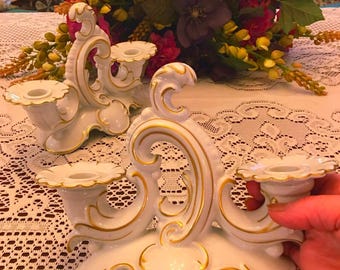 May include: Two white porcelain candelabras featuring ornate, swirling scrollwork designs and delicate gold trim along the edges. Each piece has two candle holders with scalloped rims and a central arched handle, finished with a glossy glaze.