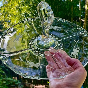 May include: A clear glass serving tray with a central handle, featuring a divided three-section design. The surface is decorated with etched floral patterns and scalloped edges, showcasing a transparent construction and a polished finish.