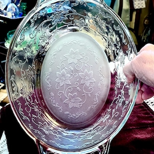 May include: Clear glass oval baking dish with floral and vine embossed detailing. The center of the dish has a frosted floral design. The dish has handles on either side. The dish is held by a hand.