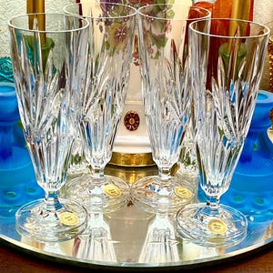Peut inclure: Quatre fl&ucirc;tes &agrave; champagne en cristal transparent avec des motifs de feuilles grav&eacute;es. Les fl&ucirc;tes sont pos&eacute;es sur une surface miroitante, avec divers vases et bougeoirs color&eacute;s en arri&egrave;re-plan. Chaque fl&ucirc;te a une base ronde avec un autocollant dor&eacute;.