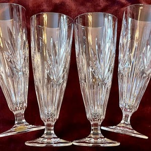 Peut inclure: Quatre fl&ucirc;tes &agrave; champagne en cristal transparent. Chaque fl&ucirc;te a un long et fin bol avec un dessus &eacute;vas&eacute; et un motif grav&eacute; d&eacute;coratif. Les fl&ucirc;tes sont pos&eacute;es sur un pied. Le fond est rouge fonc&eacute;.