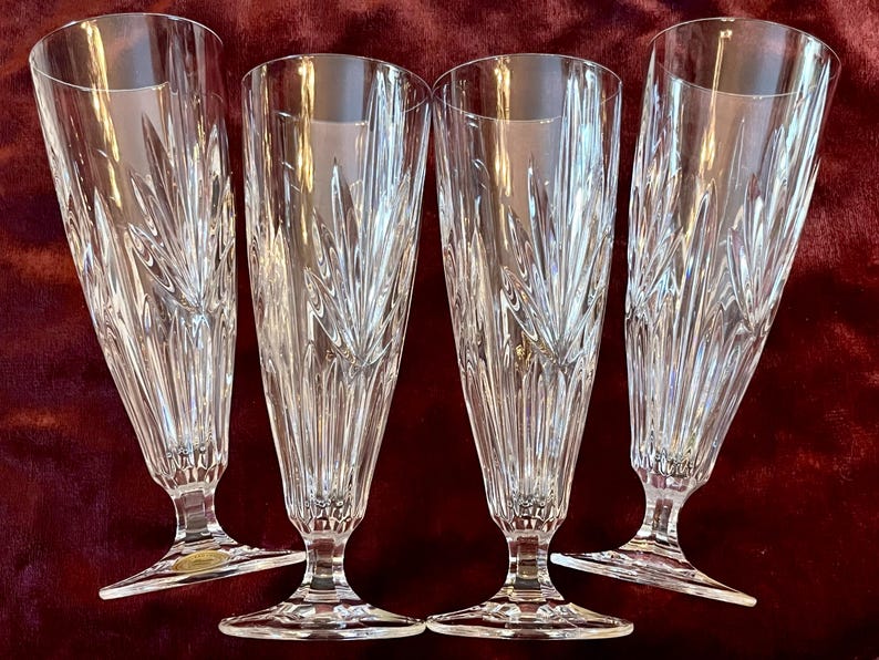 Peut inclure: Quatre fl&ucirc;tes &agrave; champagne en cristal transparent. Chaque fl&ucirc;te a un long et fin bol avec un dessus &eacute;vas&eacute; et un motif grav&eacute; d&eacute;coratif. Les fl&ucirc;tes sont pos&eacute;es sur un pied. Le fond est rouge fonc&eacute;.
