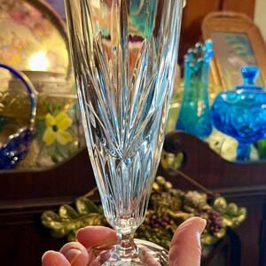 Peut inclure: Un fl&ucirc;te &agrave; champagne en cristal transparent avec un motif grav&eacute; vertical d&eacute;taill&eacute;. Le verre est tenu par une main, et une &eacute;tiquette dor&eacute;e avec du texte est visible sur la base. L'arri&egrave;re-plan comprend d'autres verreries d&eacute;coratives.