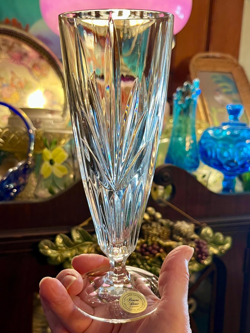Peut inclure: Un fl&ucirc;te &agrave; champagne en cristal transparent avec un motif grav&eacute; vertical d&eacute;taill&eacute;. Le verre est tenu par une main, et une &eacute;tiquette dor&eacute;e avec du texte est visible sur la base. L'arri&egrave;re-plan comprend d'autres verreries d&eacute;coratives.