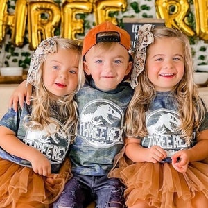 Peut inclure: Trois enfants portant des chemises assorties avec un motif de dinosaure et le texte "Three Rex" sont assis sur un banc en bois. Les enfants portent des jupes en tulle marron, des jeans et des chaussettes marron et noires. L'arrière-plan est décoré de ballons et d'une bannière en feuille d'or qui dit "Three Rex".