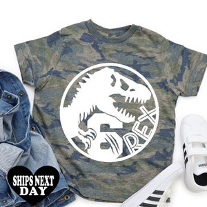 May include: Camouflage t-shirt featuring a white dinosaur graphic and the text "Rex" in a circle. The shirt is on a white background with a blue denim jacket and white trainers.