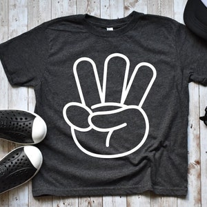 May include: A dark grey t-shirt with a white outline of a hand making the number four sign. The shirt is laid flat on a light wood background.