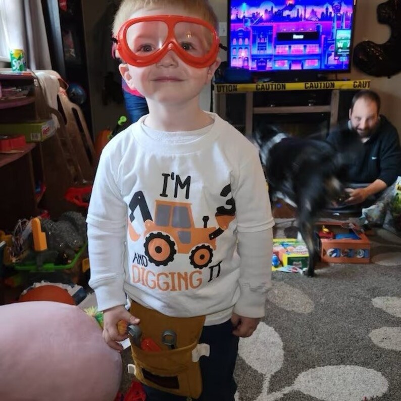 Peut inclure: Un jeune enfant portant un t-shirt blanc avec le texte "I'm 3 and digging it" et un costume de travailleur du b&acirc;timent. L'enfant porte des lunettes de s&eacute;curit&eacute; orange et tient une ceinture &agrave; outils.
