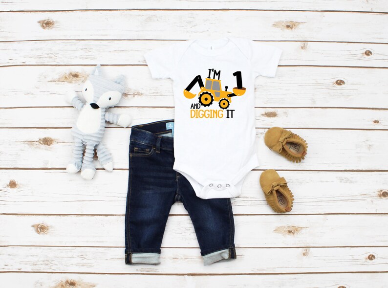 Peut inclure: Une grenouill&egrave;re blanche avec un graphique de v&eacute;hicule de chantier jaune et le texte "I'm 1 and digging it". La grenouill&egrave;re est associ&eacute;e &agrave; un jean bleu et des mocassins marrons.