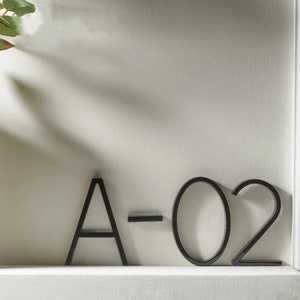 Könnte beinhalten: Schwarze Hausnummern aus Metall mit den Buchstaben A-02. Die Nummern sind an einer weißen Oberfläche befestigt.