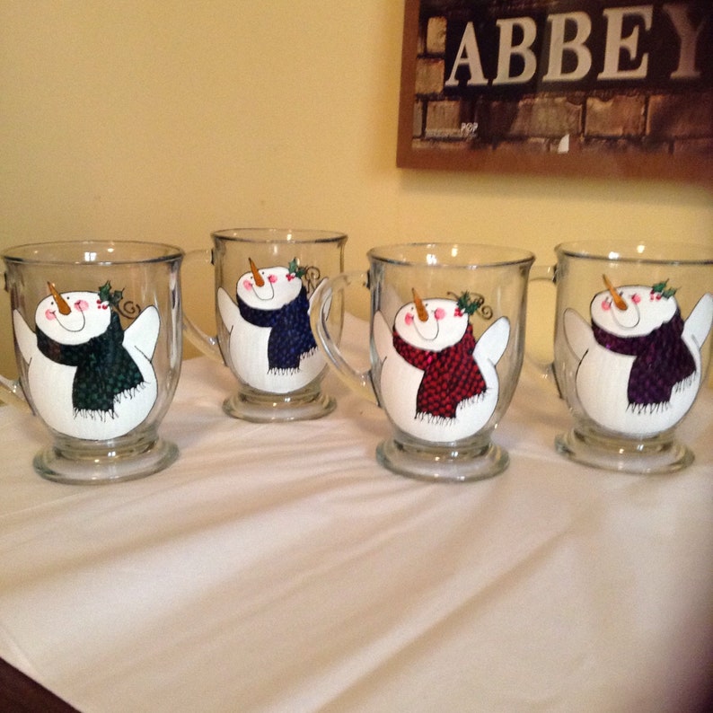 May include: Four glass mugs with snowman designs. Each mug features a snowman with a different colored scarf: green, blue, red, and purple.
