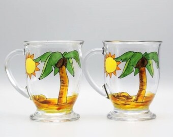 May include: Two clear glass mugs with handles, each painted with a yellow sun and a green palm tree on a brown beach.
