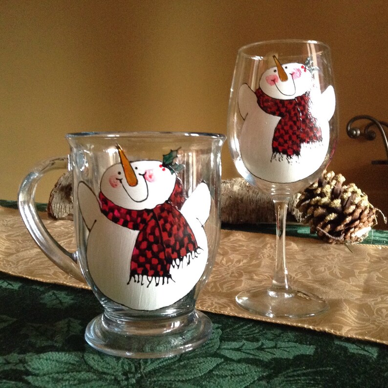 May include: Two glass mugs with hand-painted snowman designs. The snowman on the mug is wearing a red and black scarf. The snowman on the wine glass is wearing a red scarf.