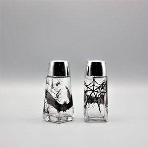 May include: A set of two glass salt and pepper shakers with silver lids. The shakers are decorated with black and white Halloween-themed designs, including bats, spiders, and spiderwebs.