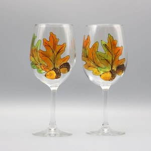 May include: Two clear glass wine glasses with hand-painted autumn leaves and acorns. The leaves are shades of orange, yellow, and green.