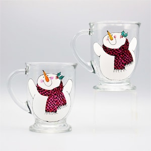 May include: Two glass mugs with snowman designs. Each mug features a white snowman with a pink and white checkered scarf, a black hat, and a carrot nose. One snowman has a green holly sprig on its hat.