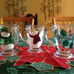 May include: Three glass mugs with painted holiday designs. The mugs feature a snowman, a snowman with a red scarf, and holly with red berries.