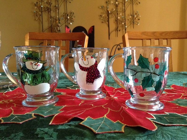 May include: Three glass mugs with painted holiday designs. The mugs feature a snowman, a snowman with a red scarf, and holly with red berries.