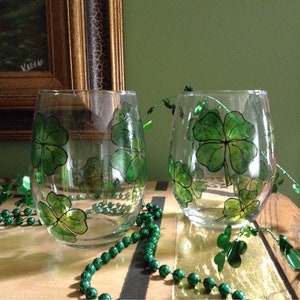 May include: Two clear glass wine glasses with green shamrock designs painted on them. The glasses are sitting on a wooden surface with green beads scattered around them.
