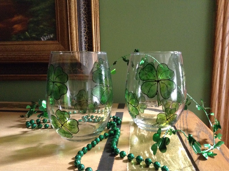 May include: Two clear glass wine glasses with green shamrock designs painted on them. The glasses are sitting on a wooden surface with green beads scattered around them.