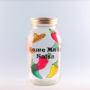 May include: A clear glass jar with a gold lid, decorated with hand-painted red, yellow, and green chili peppers and a yellow sombrero. The jar is labeled "Home Made Salsa".