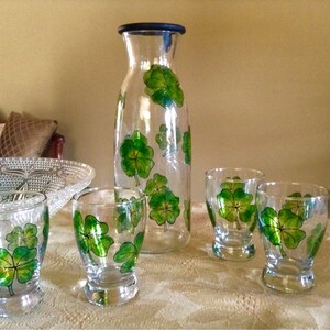 May include: A set of four clear glass drinking glasses and a matching glass pitcher, all decorated with hand-painted green four-leaf clover designs.
